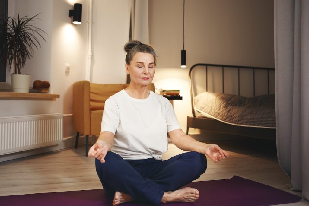 Seniorin sitzt in ihrem Wohnzimmer auf dem Boden und meditiert