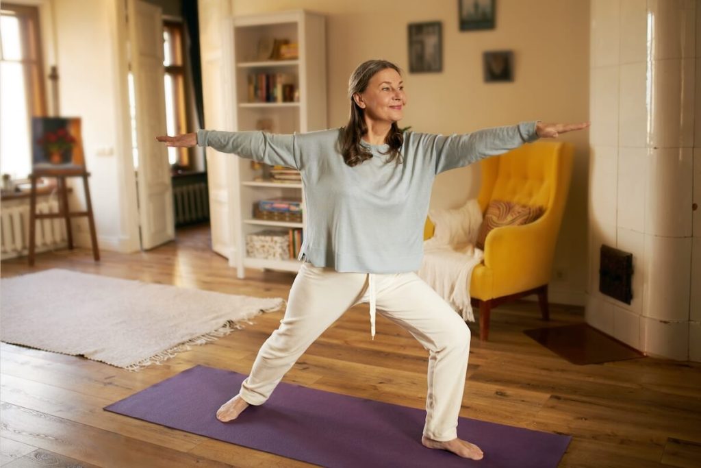 Eine Seniorin praktiziert Achtsames Yoga in ihrem Wohnzimmer